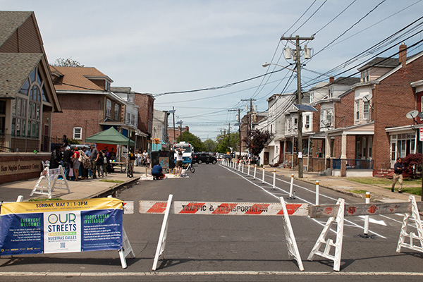 View of a residential street with a protected bike lane and a tent with people, a sign and barricade in the foreground. Credit Clyde Scott. 