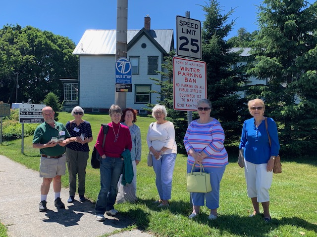 A group of people standing by a bus stop sign with a house in the background. Credit: Mpact